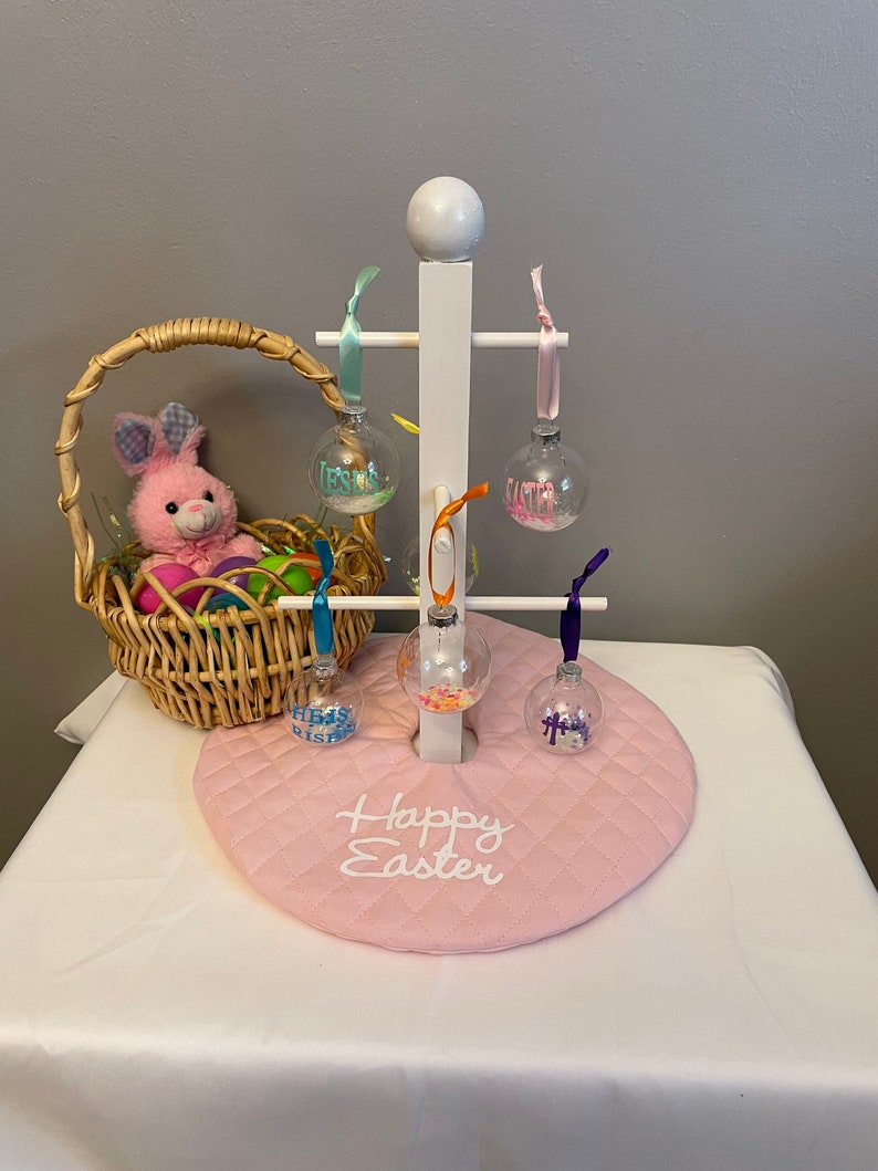 May include: A white wooden stand with four clear glass ornaments hanging from it. The ornaments are filled with colorful confetti and have the words "Happy Easter" written on them. The stand is sitting on a pink quilted mat with the words "Happy Easter" embroidered on it.