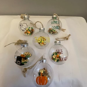 May include: Six clear glass ornaments with silver tops and twine hangers. Each ornament contains colorful sequins and a Thanksgiving-themed design, including a turkey, pumpkin, and the phrases "Gobble Gobble" and "Give Thanks."