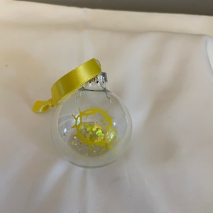 May include: A clear glass Christmas ornament with a yellow glitter crown of thorns inside. The ornament is decorated with a yellow ribbon.