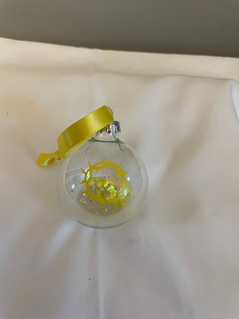 May include: A clear glass Christmas ornament with a yellow glitter crown of thorns inside. The ornament is decorated with a yellow ribbon.