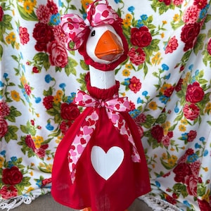 May include: A white goose figurine wearing a red dress with pink hearts and a pink bow. The goose is standing on a white background with a floral pattern.