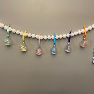 May include: A white pom-pom garland with seven glass ornaments hanging from it. The ornaments are filled with glitter and have different colored ribbons and words on them, including "Jesus", "Faith", "Hope", "Love", and "Joy".
