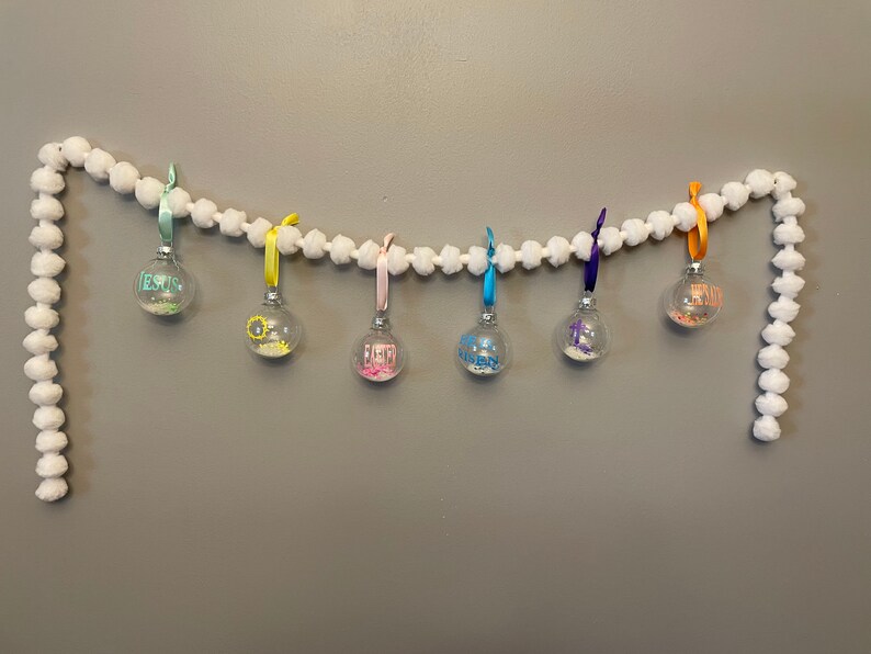 May include: A white pom-pom garland with seven glass ornaments hanging from it. The ornaments are filled with glitter and have different colored ribbons and words on them, including "Jesus", "Faith", "Hope", "Love", and "Joy".
