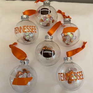 May include: Five clear glass Christmas ornaments with orange and white ribbons. The ornaments are filled with orange and iridescent sequins.  Three of the ornaments have orange vinyl designs: a football, the state of Tennessee, and the word "Tennessee".