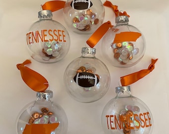 May include: Five clear glass Christmas ornaments with orange and white ribbons. The ornaments are filled with orange and iridescent sequins.  Three of the ornaments have orange vinyl designs: a football, the state of Tennessee, and the word "Tennessee".