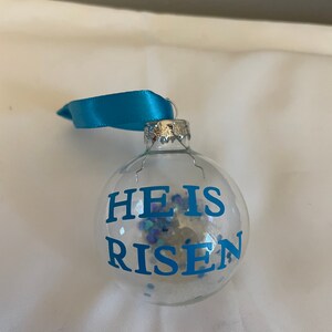 May include: A clear glass Christmas ornament with a blue ribbon. The ornament has blue glitter inside and the words "He is Risen" written in blue on the front.