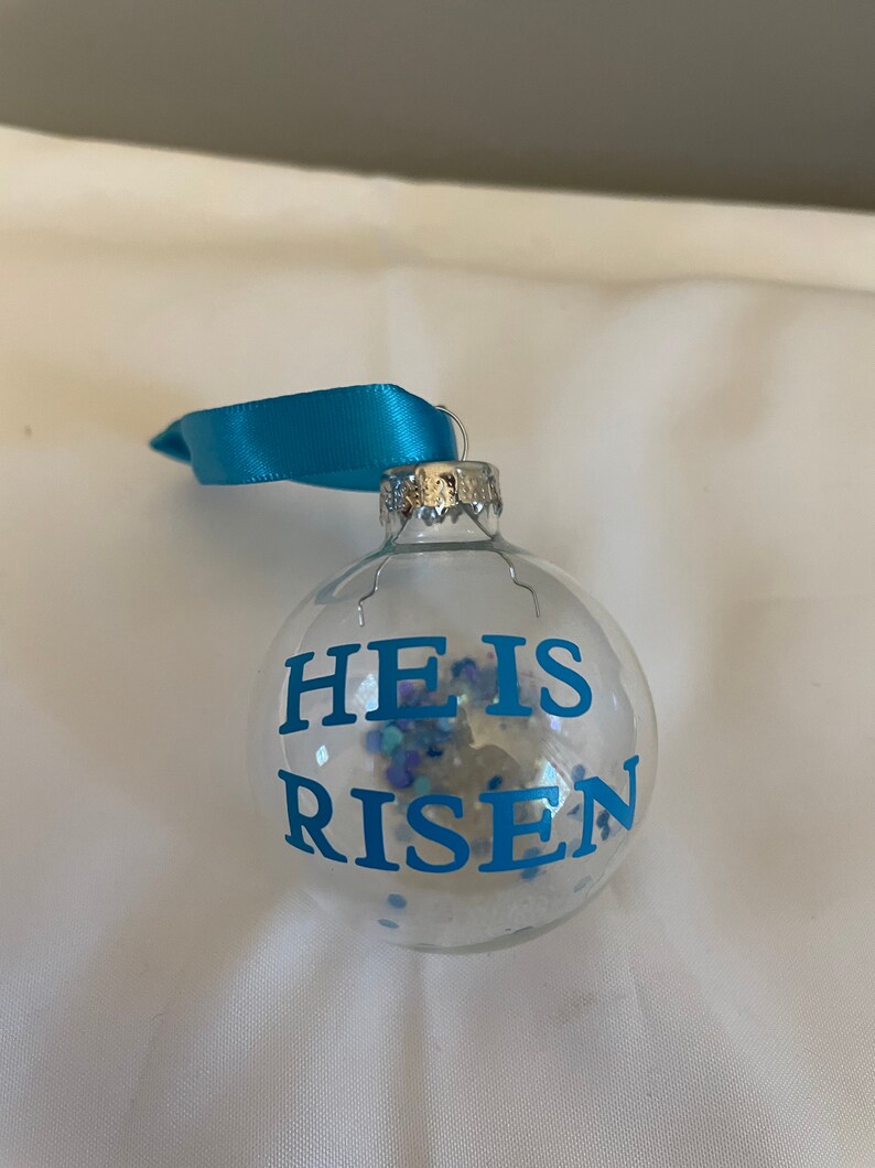 May include: A clear glass Christmas ornament with a blue ribbon. The ornament has blue glitter inside and the words "He is Risen" written in blue on the front.