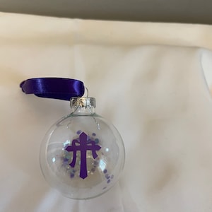 May include: A clear glass Christmas ornament with a purple cross and purple and white glitter inside. The ornament has a purple ribbon attached to the top.