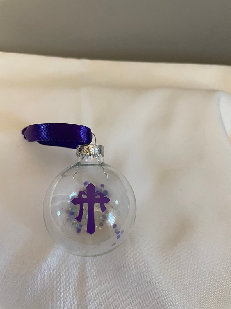 May include: A clear glass Christmas ornament with a purple cross and purple and white glitter inside. The ornament has a purple ribbon attached to the top.
