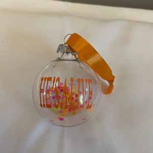 May include: A clear glass Christmas ornament with an orange ribbon and the text "HE'S ALIVE" in orange lettering. The ornament is filled with colorful glitter.