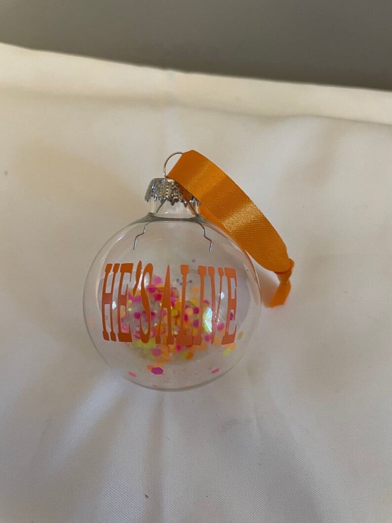 May include: A clear glass Christmas ornament with an orange ribbon and the text "HE'S ALIVE" in orange lettering. The ornament is filled with colorful glitter.