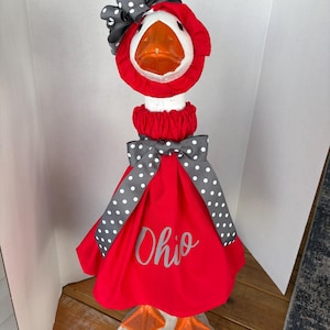 May include: A decorative goose statue wearing a red dress with the word "Ohio" in silver script. The goose has a white head, orange beak and feet, and is adorned with a gray and white polka dot bow and hat.