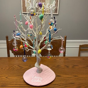 May include: A white twig tree decorated with clear glass ornaments filled with colorful Easter eggs. The tree is on a pink table runner with the words "Happy Easter" embroidered on it. A wooden sign with the word "Blessed" is hanging on the wall behind the tree.