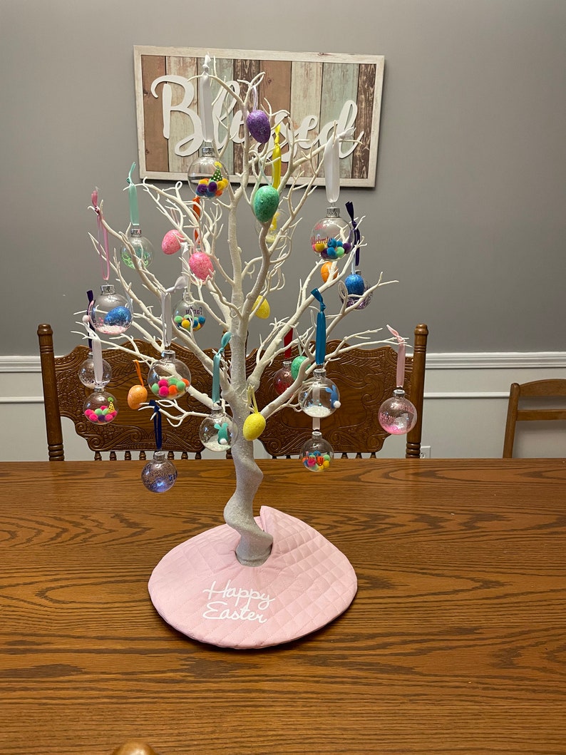 May include: A white twig tree decorated with clear glass ornaments filled with colorful Easter eggs. The tree is on a pink table runner with the words "Happy Easter" embroidered on it. A wooden sign with the word "Blessed" is hanging on the wall behind the tree.