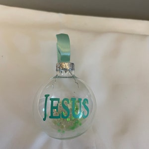 May include: A clear glass ornament with a green ribbon and the word "JESUS" in green lettering. The ornament is filled with green glitter.