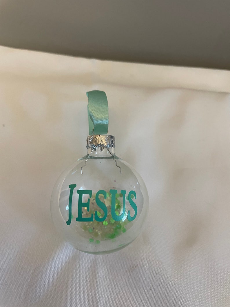 May include: A clear glass ornament with a green ribbon and the word "JESUS" in green lettering. The ornament is filled with green glitter.