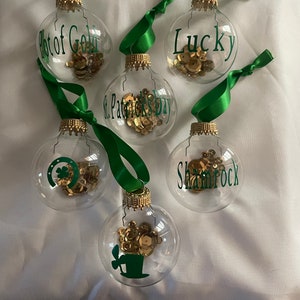 May include: Five clear glass ornaments with gold glitter inside and green ribbon. The ornaments are decorated with green vinyl lettering that reads "Pot of Gold", "Lucky", "St. Patrick's Day", "Shamrock", and a leprechaun hat.