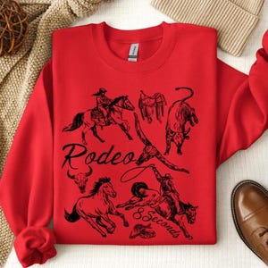 May include: A red sweatshirt featuring black illustrations of rodeo events, including riders on horses, a bull, and a saddle. The word "Rodeo" is written in a script font, with "8 Seconds" below.