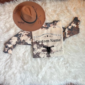 Op de afbeelding: Een bruin en wit tie-dye sweatshirt met een zwart-witte afbeelding van een koeien schedel in een rechthoek met de tekst "Custom Name".
