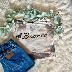 May include: A light brown t-shirt with a tie-dye pattern. The shirt has a black silhouette of a horse with the word "Bronco" in gold lettering.