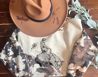 May include: A white sweatshirt with brown and black tie-dye pattern on the sleeves. The front of the sweatshirt features a black and white illustration of a cowboy riding a horse. The text "Roo" is written on a brown hat in the background.