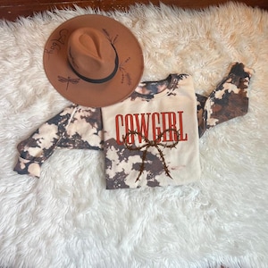 May include: A brown fedora hat with a black band and decorative dragonfly and floral designs, next to a long-sleeved shirt. The shirt is cream-colored with a brown and white pattern and the word "COWGIRL" in red.