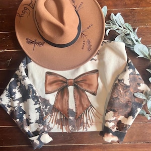 May include: A white sweatshirt with brown splatter paint and a brown bow with fringe. The bow has a stitched design. A brown hat with a dragonfly design is on top of the sweatshirt.