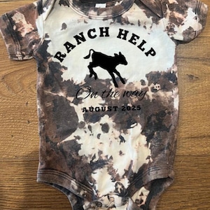 Könnte beinhalten: Kurzärmeliger Baby-Body mit braunem und weißem Batikmuster. Der Body zeigt den Text "RANCH HELP" über einer schwarzen Silhouette eines Kalbes, mit "On the way" und "AUGUST 2025" darunter.
