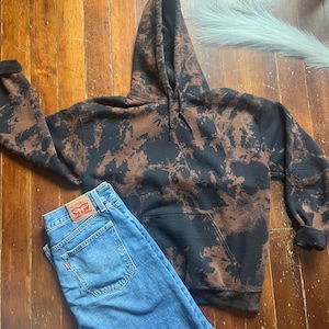 Könnte beinhalten: Ein schwarzer Kapuzenpullover mit braunen Bleichflecken und einer Vordertasche wird mit einer blauen Jeans kombiniert. Die Jeans hat ein rotes Levi's-Etikett auf der Gesäßtasche. Der Kapuzenpullover hat eine Kapuze mit Kordelzug und gerippte Bündchen.