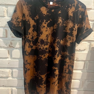 Puede incluir: Camiseta negra con un diseño tie-dye marrón. La camiseta de manga corta está colgada en una percha de madera contra una pared de ladrillos blancos. Las mangas están dobladas.