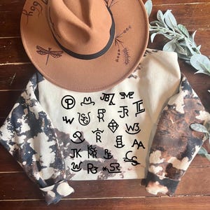 May include: A white sweatshirt with brown splatter paint and black brand marks. The sweatshirt has long sleeves and a crew neck. The brand marks are in a variety of styles, including circles, squares, and diamonds.