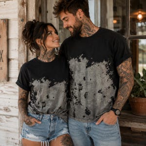 May include: Two black t-shirts with a distressed, bleached-effect gray pattern across the midsection. The shirts feature short sleeves with rolled cuffs and a classic crew neckline, showcasing a faded, mottled texture that transitions from black to gray.