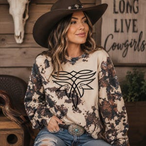 May include: A long-sleeve crewneck sweatshirt featuring a beige base with a dark brown and black acid-wash pattern. The center of the chest displays a black, symmetrical, linear graphic design composed of interconnected, curved, and pointed shapes.