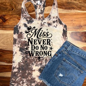 Op de afbeelding: Een tie-dye tanktop met de tekst "Miss Never Do No Wrong" in zwart schrift en een cowboyhoed-afbeelding. Gecombineerd met een spijkerbroekshort. De tanktop heeft een bruin en crèmekleurig patroon.