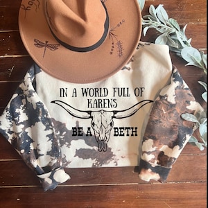 May include: A tan fedora hat with a black band and dragonfly and floral designs sits atop a cream-colored sweatshirt. The sweatshirt features a cow skull graphic and the text "IN A WORLD FULL OF KARENS BE A BETH". The sleeves have a unique bleached pattern.