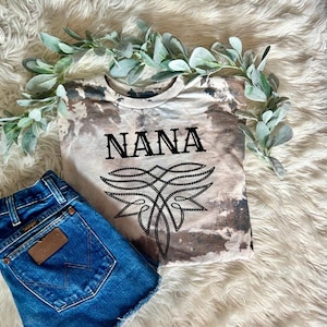 May include: A light brown t-shirt with a tie-dye pattern and the word "NANA" in black lettering. The shirt has a black and white design underneath the word "NANA".