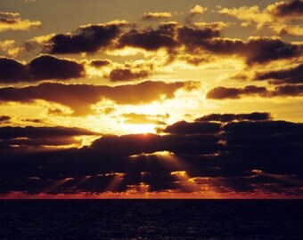 May include: A dramatic sunset over a dark body of water with a golden sky and dark clouds. The sun is hidden behind the clouds, but its rays are visible through the clouds.