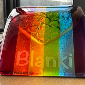 Puede incluir: Un monumento conmemorativo para mascotas de vidrio rectangular con un esquema de color arco&iacute;ris. El monumento conmemora una huella de pata y el nombre "Blanki" grabado. Los colores van del rojo al morado.
