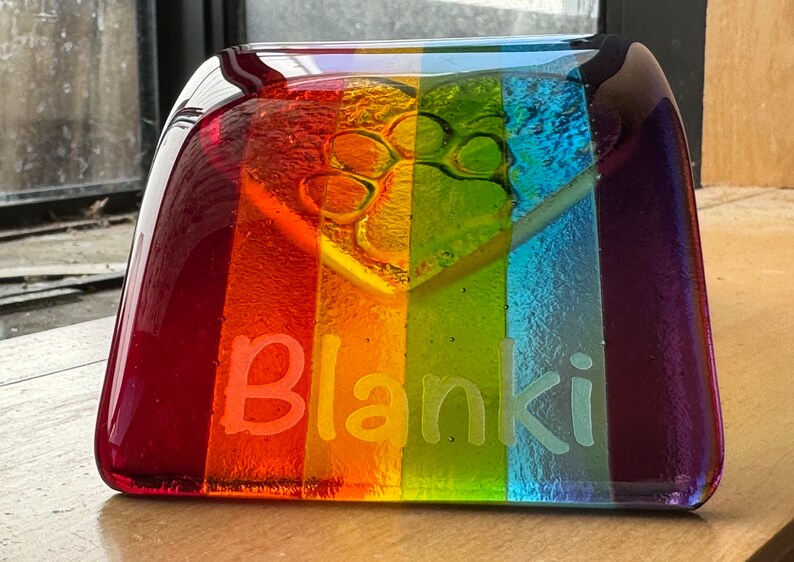 Puede incluir: Un monumento conmemorativo para mascotas de vidrio rectangular con un esquema de color arco&iacute;ris. El monumento conmemora una huella de pata y el nombre "Blanki" grabado. Los colores van del rojo al morado.