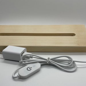 Puede incluir: Una base de madera blanca con una ranura larga y estrecha en el centro. Un adaptador de corriente blanco y un cable blanco con un interruptor de encendido/apagado están conectados a la base.