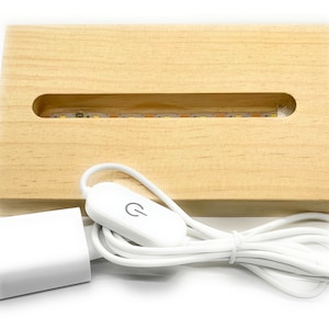 Puede incluir: Una base de madera con un hueco para exhibir objetos, que cuenta con una tira de luz LED integrada. La base está conectada a un adaptador de corriente blanco con un interruptor.