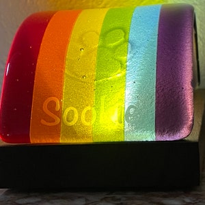Puede incluir: Una base de lámpara de vidrio con colores de arcoíris con la palabra "Sophie" grabada en ella. La base está iluminada desde adentro, proyectando un brillo cálido. Se asienta sobre una base de madera negra.