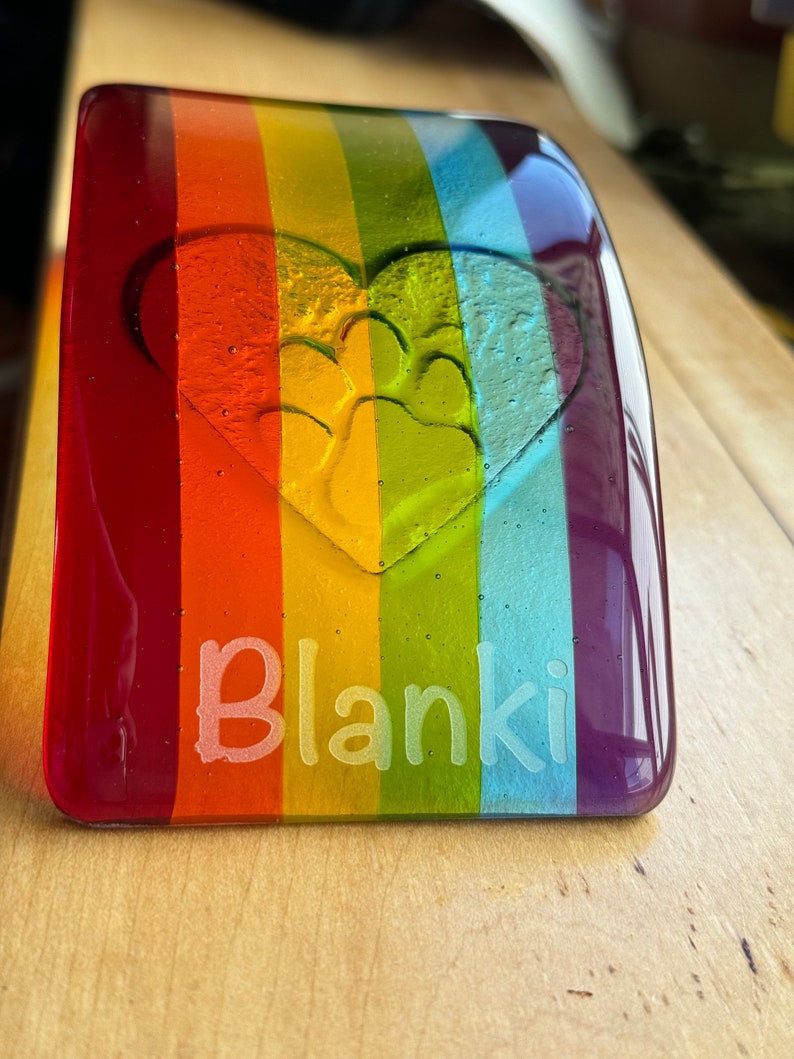 Puede incluir: Un memorial para mascotas de vidrio rectangular con un dise&ntilde;o de arco&iacute;ris. El vidrio presenta un coraz&oacute;n con una huella de pata y el nombre "Blanki" grabado en blanco. Los colores incluyen rojo, naranja, amarillo, verde, azul y morado.