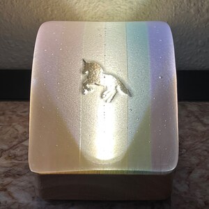 Puede incluir: Una luz nocturna blanca con un diseño de arcoíris y una silueta de unicornio dorada. La luz está encendida e ilumina el unicornio.