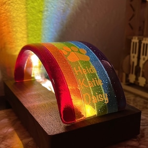 Puede incluir: Un arco de cristal de colores arcoíris con purpurina incrustada en el cristal. El arco está iluminado desde el interior y tiene los nombres "Heidi", "Katie" y "Daisy" grabados en él. También hay una huella de pata grabada en el arco.