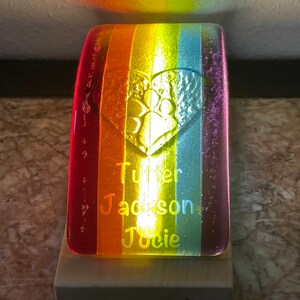 Puede incluir: Placa de vidrio rectangular con los colores del arco&iacute;ris, con un dise&ntilde;o de huella en forma de coraz&oacute;n. Los nombres "Tutter", "Jackson" y "Jocie" est&aacute;n grabados debajo. La placa est&aacute; montada sobre una base de madera.
