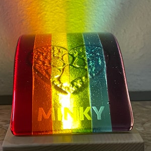 Puede incluir: Una colorida pieza de arte en vidrio con un dise&ntilde;o de arco&iacute;ris. La pieza presenta un dise&ntilde;o de &aacute;rbol en el centro y la palabra "MINKY" en la parte inferior. El vidrio est&aacute; montado sobre una base de madera.