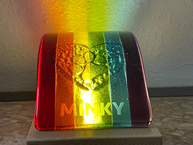 Puede incluir: Una colorida pieza de arte en vidrio con un dise&ntilde;o de arco&iacute;ris. La pieza presenta un dise&ntilde;o de &aacute;rbol en el centro y la palabra "MINKY" en la parte inferior. El vidrio est&aacute; montado sobre una base de madera.