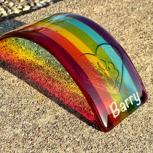 Puede incluir: Una pieza de arte de vidrio curvada, con los colores del arco&iacute;ris, con un dise&ntilde;o de huella de pata y el nombre "Barry" en blanco. El vidrio presenta un degradado de colores, incluyendo rojo, naranja, amarillo, verde, azul y morado.