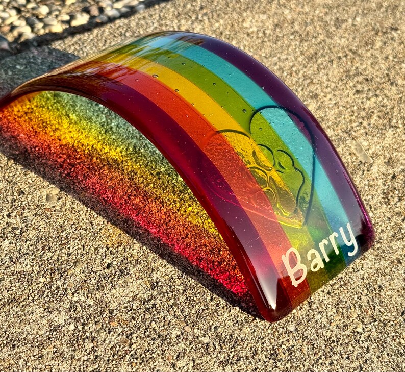 Puede incluir: Una pieza de arte de vidrio curvada, con los colores del arco&iacute;ris, con un dise&ntilde;o de huella de pata y el nombre "Barry" en blanco. El vidrio presenta un degradado de colores, incluyendo rojo, naranja, amarillo, verde, azul y morado.